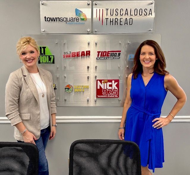 Two women smiling in a radio studio with station logos on the wall