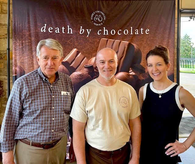 Three people posing in front of a “death by chocolate” banner and stacked chocolate bars.