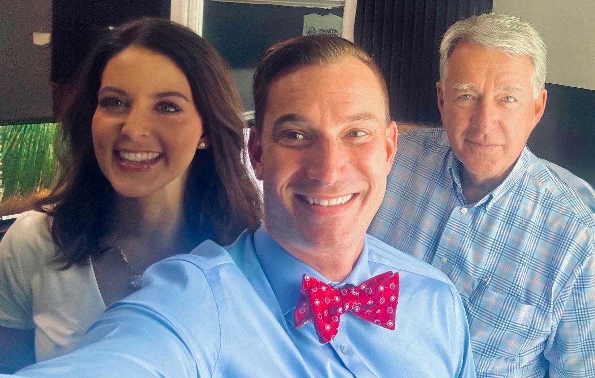 Three smiling people posing indoors, with the front man wearing a red polka-dot bow tie.