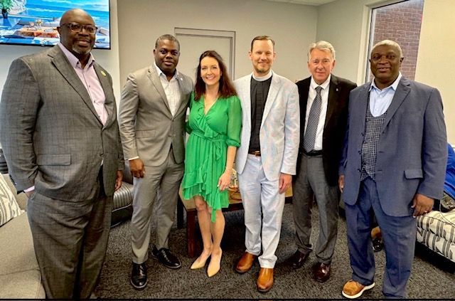 Five people in business attire stand smiling in an office setting.