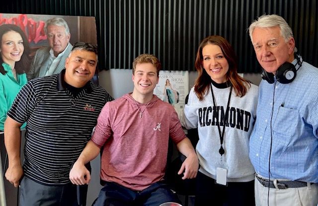 Four people smiling in a studio, with one seated and an older man wearing headphones standing beside them.