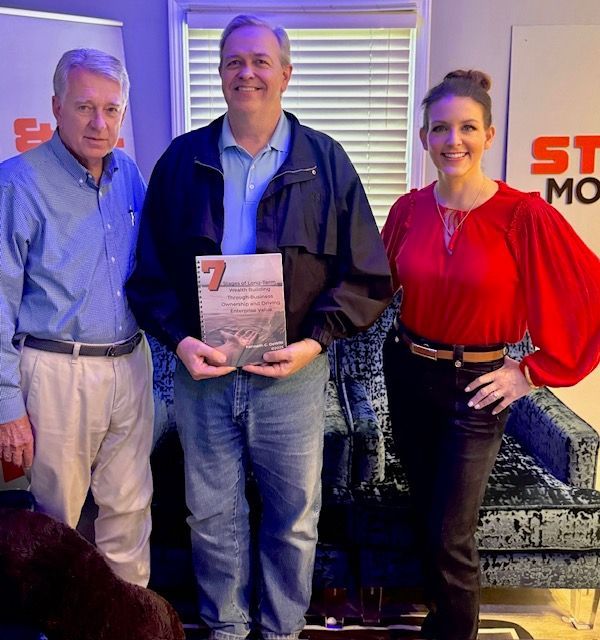 Three people pose indoors, one holding an award plaque, with a couch and window blinds behind them.