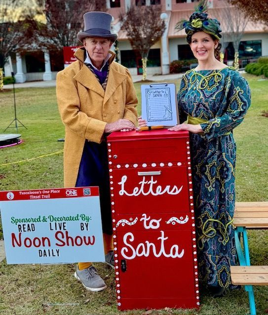 Two people stand by a red “Letters to Santa” box and a “Noon Show Daily” sign outdoors.
