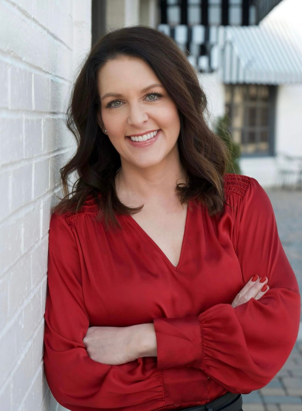 Woman in a red blouse smiling with arms crossed against a light wall outdoors