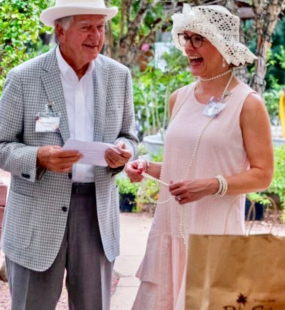 Two people in light summer clothes chatting outdoors, one holding a paper and the other a small item.