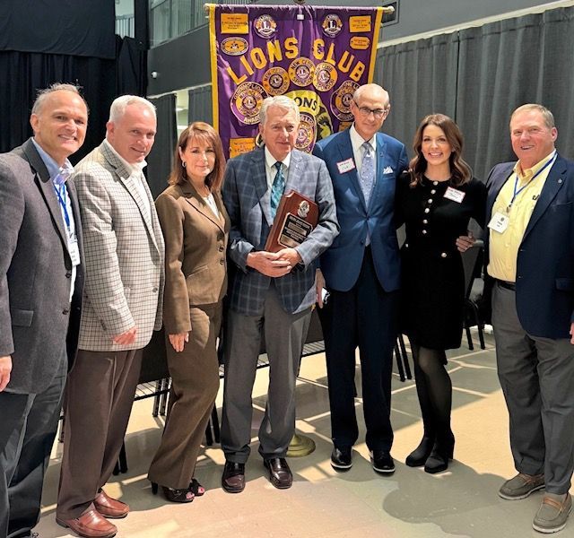 Group of seven people standing on a stage, posing with a Lions Club banner; one man holds a plaque.
