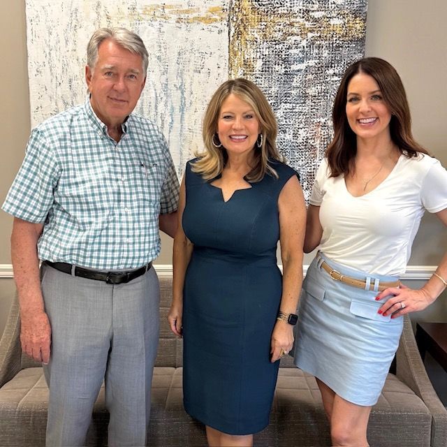 Three people standing and smiling in an office lounge beside a gray couch and abstract wall art.