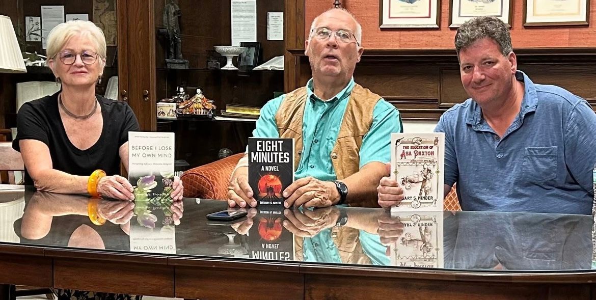 Three people seated behind a table with three books displayed in a library or office setting