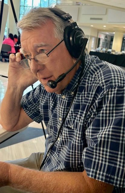 Man wearing a headset and plaid shirt, seated indoors at a desk, resting his head on his hand.