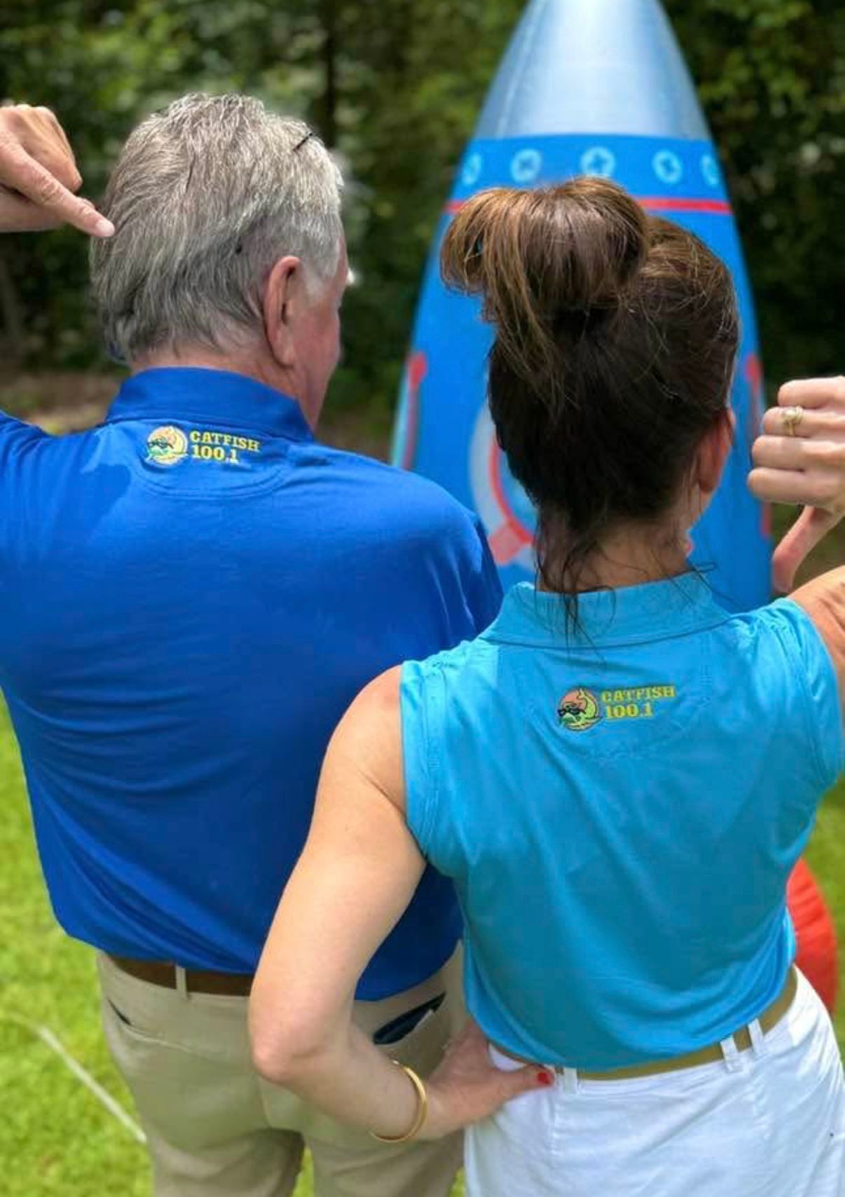 Two people in blue shirts stand outdoors facing a blue event marker, seen from behind