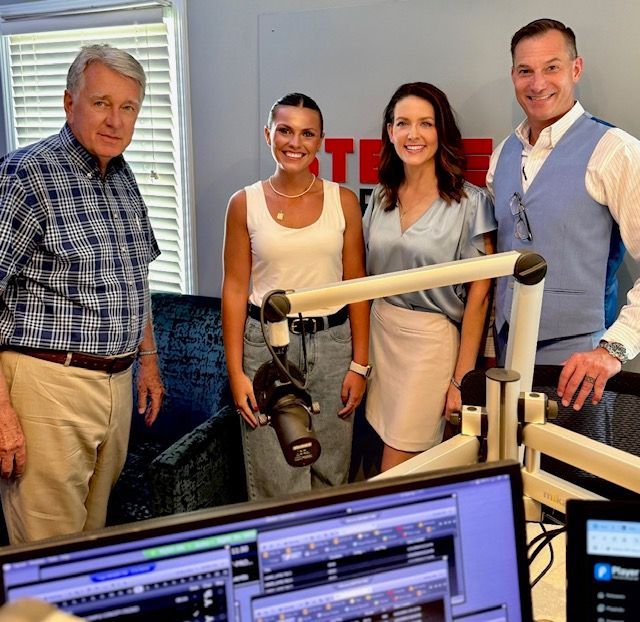 Four people standing in a radio studio behind microphones and a mixing console