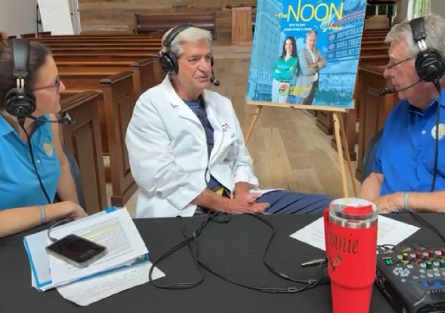 Three podcast hosts wearing headsets record at a table in a church pew setting.