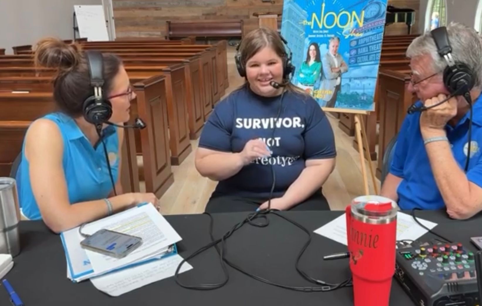 Three podcast hosts wearing headsets at a table, speaking into microphones in a wood-paneled room