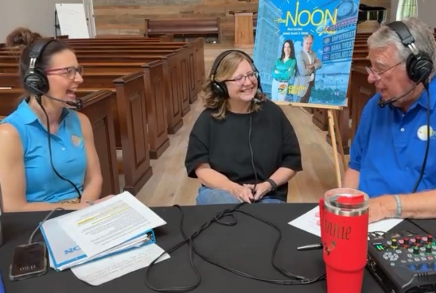 Three people wearing headsets record a talk show at a table with papers and a mixer.