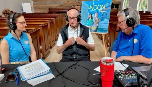 Three people wearing headphones record a podcast at a table, with a blue “Noon” poster behind them.