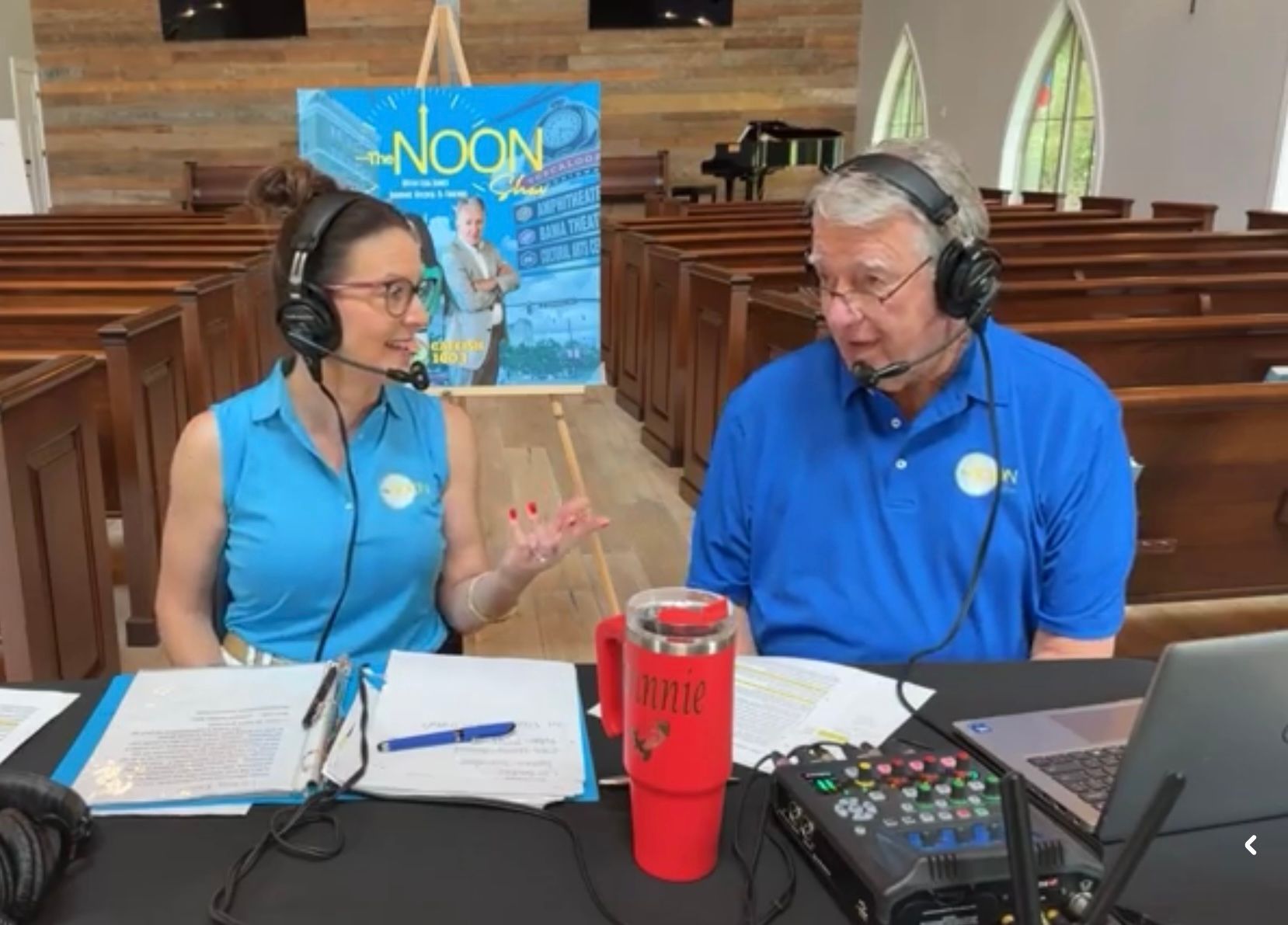 Two people wearing headsets record a podcast at a table in a church auditorium.