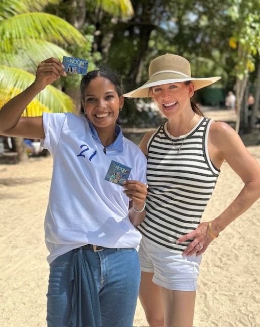 Two smiling people posing outdoors, holding small cards, one in a blue shirt and jeans, the other in a striped top and sunhat.