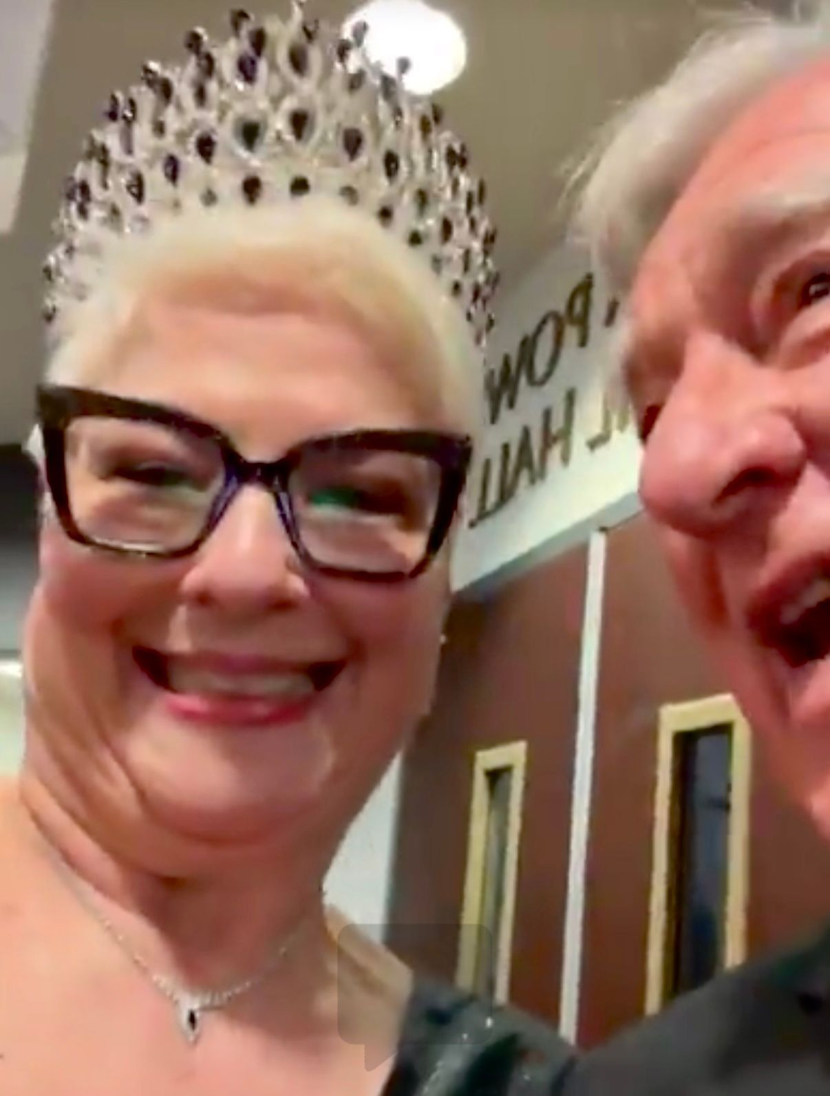 Two smiling people indoors, one wearing glasses and a jeweled crown, in front of a door with a sign.