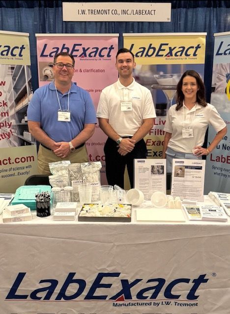 Three people at a LabExact trade show booth with product displays and branded banners