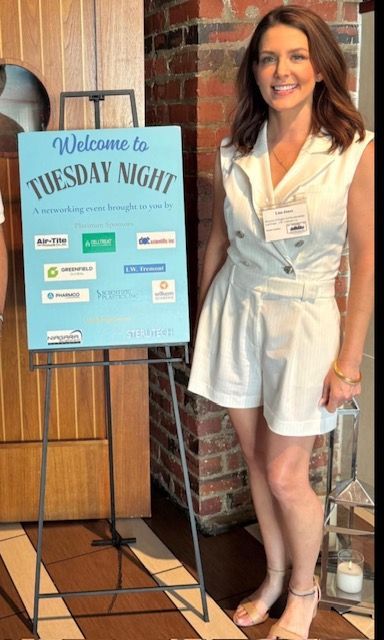 Woman standing beside a blue “Welcome to Tuesday Night” sign in a brick entryway.