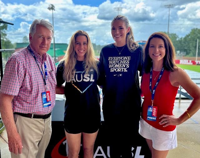 Four people posing outdoors at a sporting event, two wearing athletic uniforms with sponsor logos.