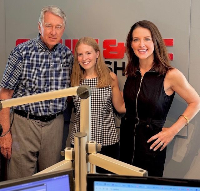 Three people pose in a radio studio, smiling behind microphones and equipment.