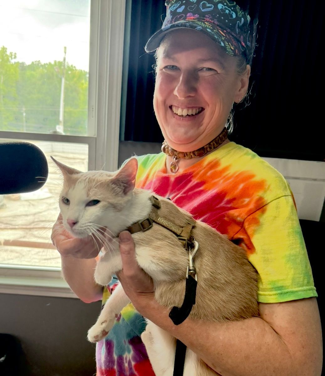 Smiling person in tie-dye shirt holding a white cat indoors near a window