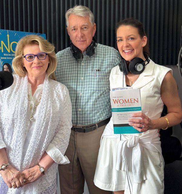 Three people posing in a studio; one holds a book titled “WOMEN,” with headphones around her neck.