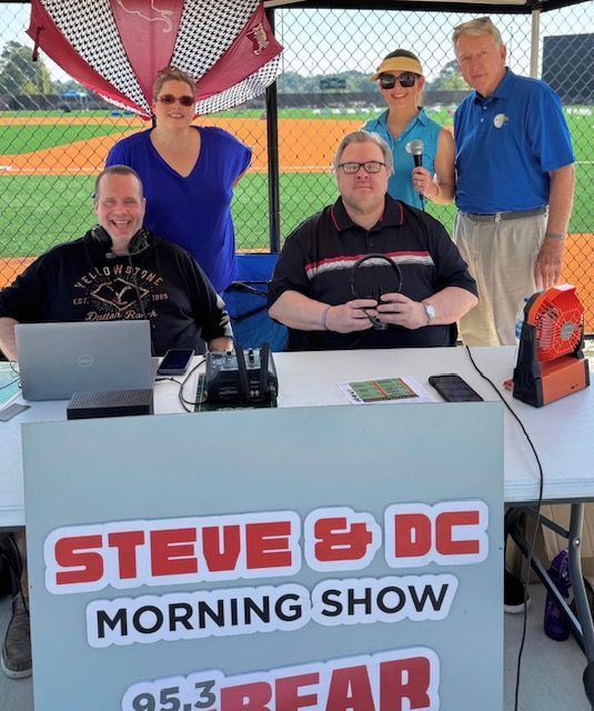 Radio show booth with five people behind a table labeled “STEVE & DC MORNING SHOW” at a ballpark