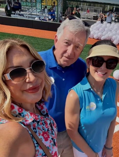 Three people smiling at a baseball field on a sunny day, with seats and field visible behind them
