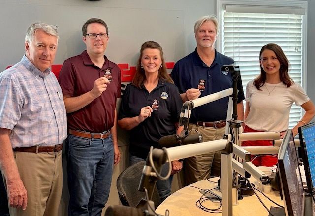 Five people posing in a radio studio with microphones and equipment, smiling at the camera.