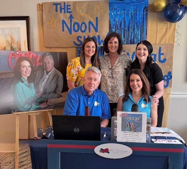 Booth with five smiling people behind a table, banner reading “The Noontime Show,” and a laptop display