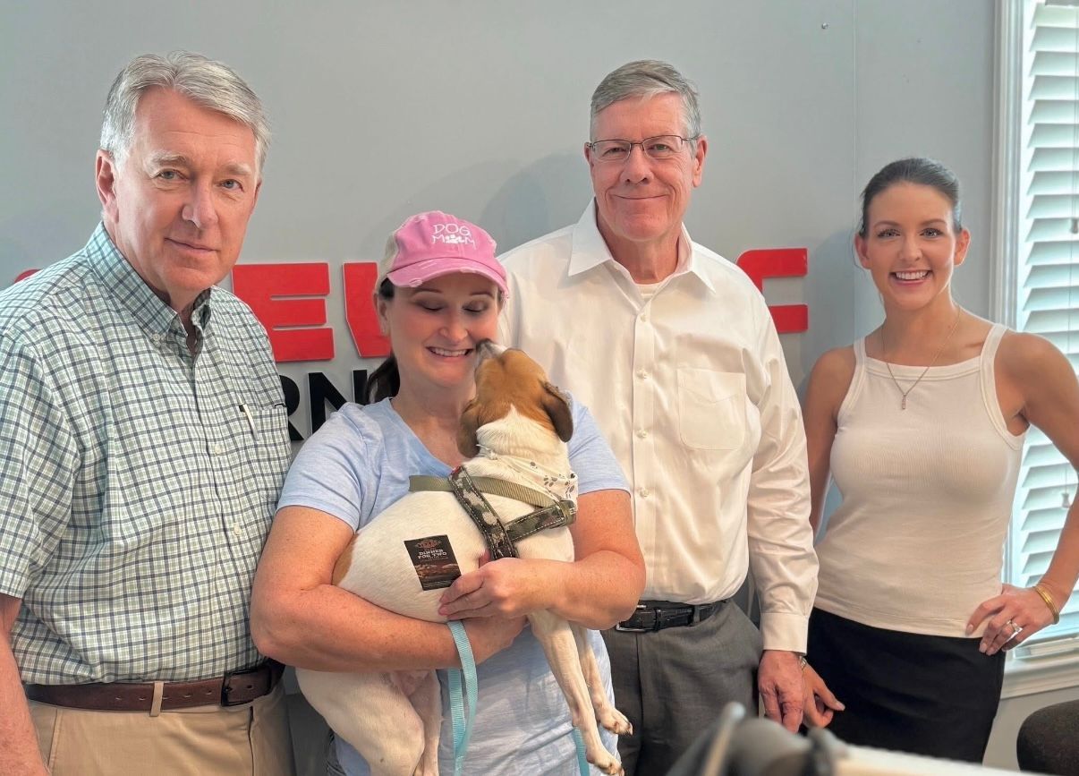 Four people posing indoors, one holding a small dog in front of a red wall sign.
