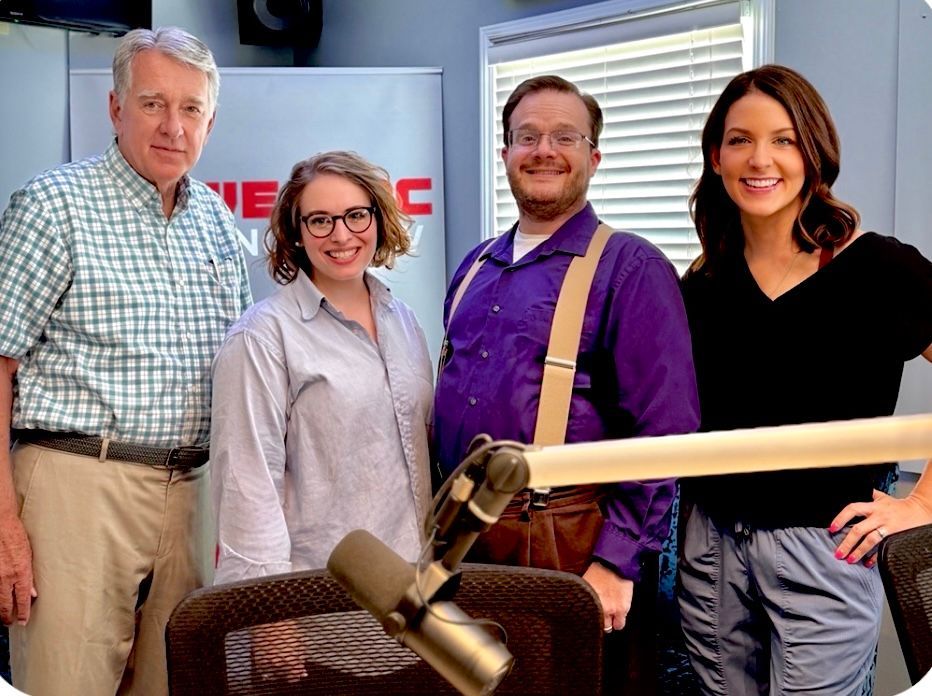 Four people posing in a radio studio with microphones and a desk in front of them.