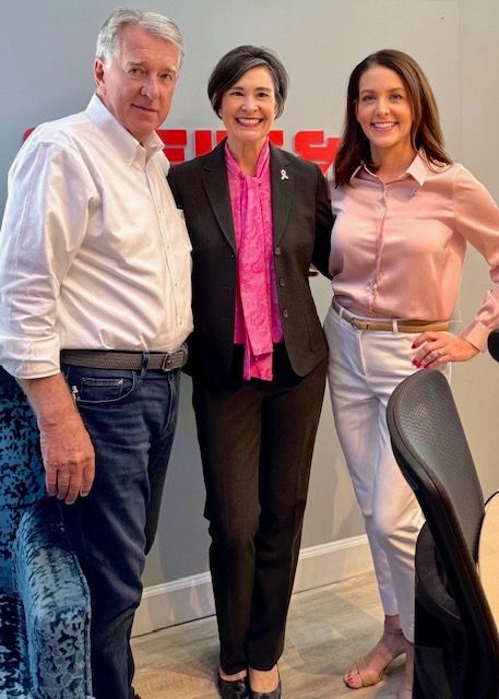 Three people posing indoors, smiling beside a desk chair.