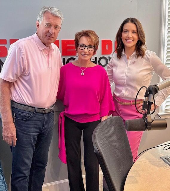 Three people posing in a radio studio, with microphones and a branded backdrop.