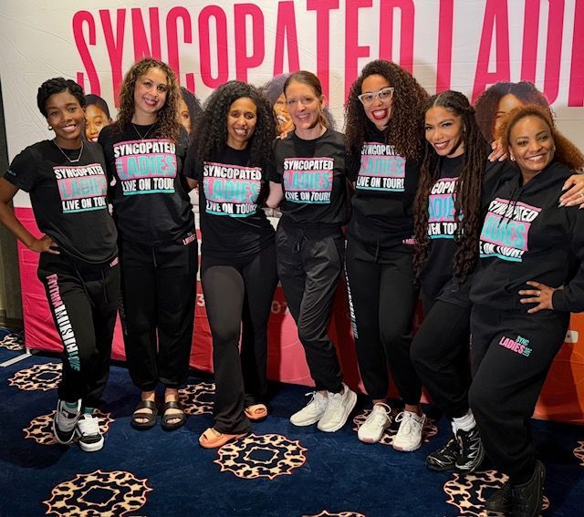 Seven people posing in front of a SYNCOPATED backdrop, wearing matching black T-shirts and jeans.
