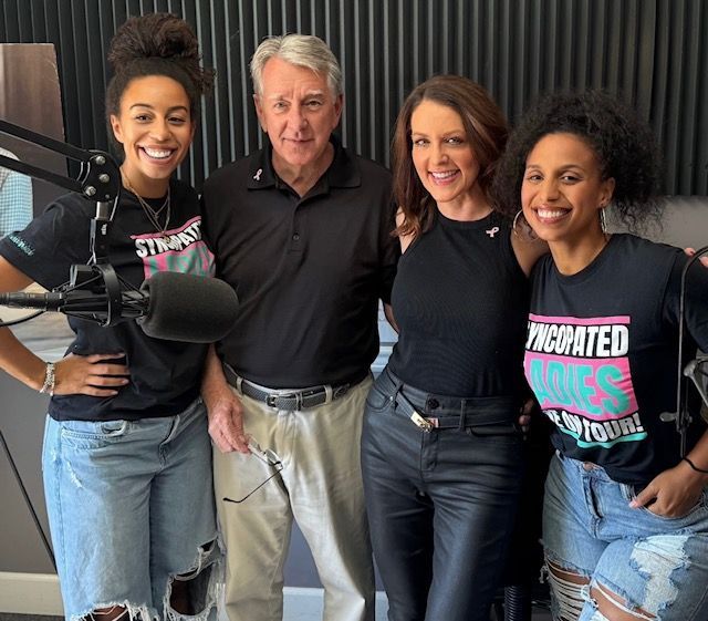 Four people standing together in a radio studio, smiling beside a microphone.