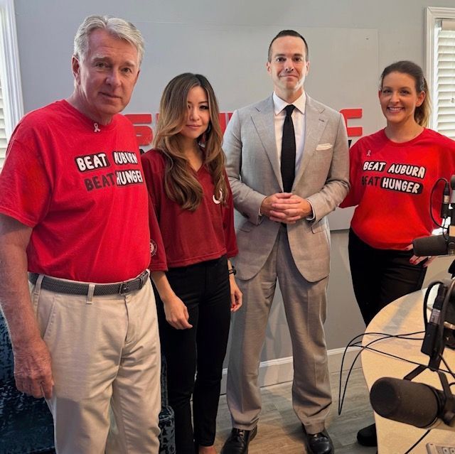 Four people standing in a studio, two wearing red “Beat Hunger” shirts beside a man in a suit.