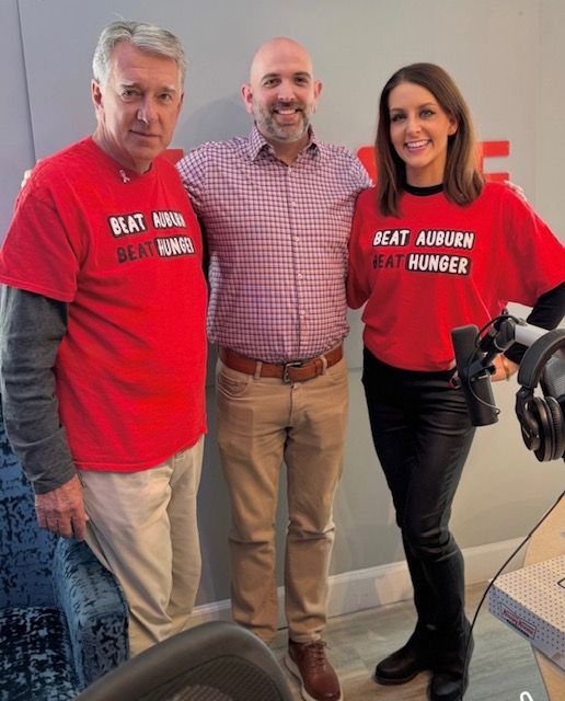 Three adults standing indoors, wearing red hunger awareness shirts and smiling at the camera.