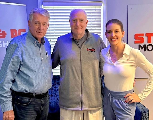 Three people posing indoors in front of a branded backdrop, smiling at the camera.