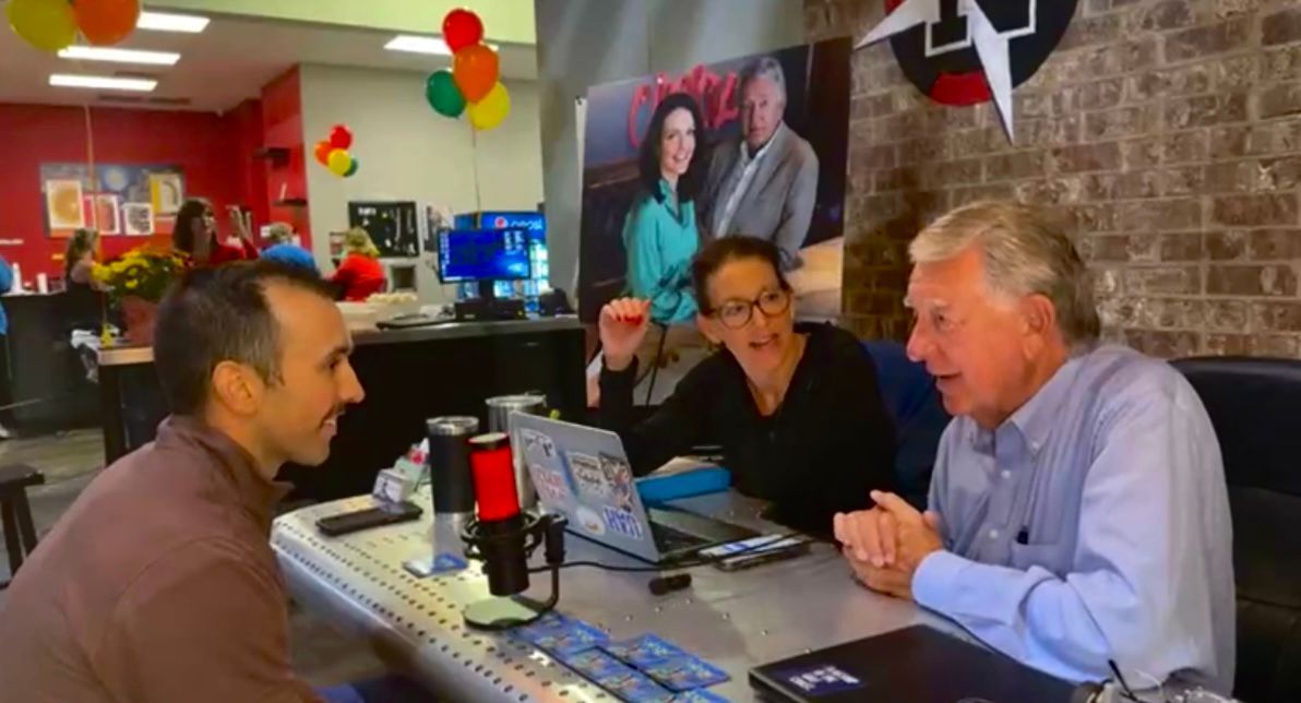 Three people chatting at a table in a colorful café or shop, with balloons and a poster in the background