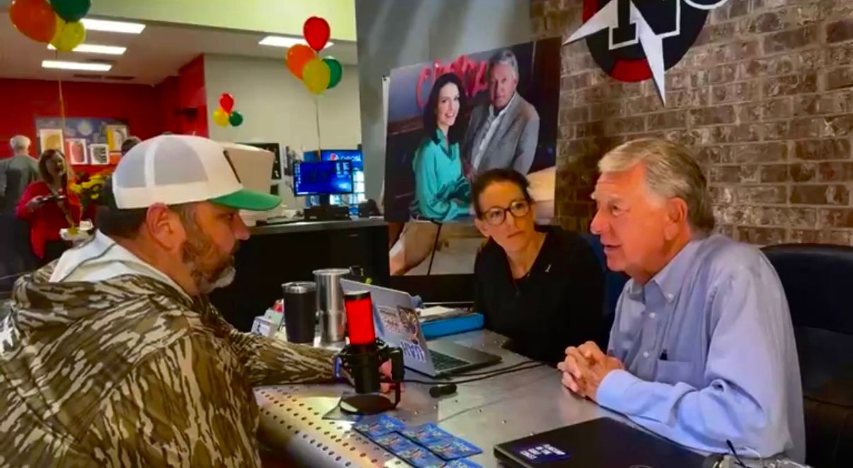 Three men in a colorful radio studio, talking across a desk with microphones and monitors.