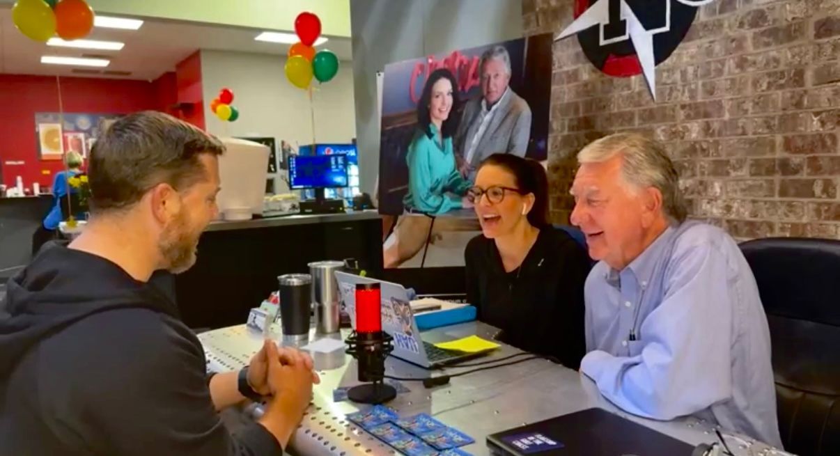 Three people chatting at a desk in a festive office with balloons and a wall poster.
