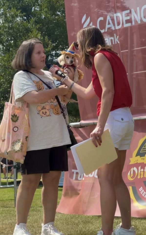 Two people exchanging a small dog on a leash at an outdoor event booth with banners.