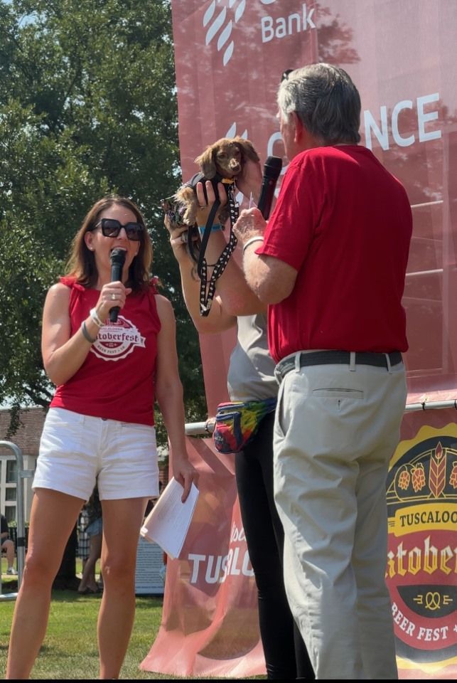 Three people on stage at an outdoor event, speaking into microphones in front of a branded banner.