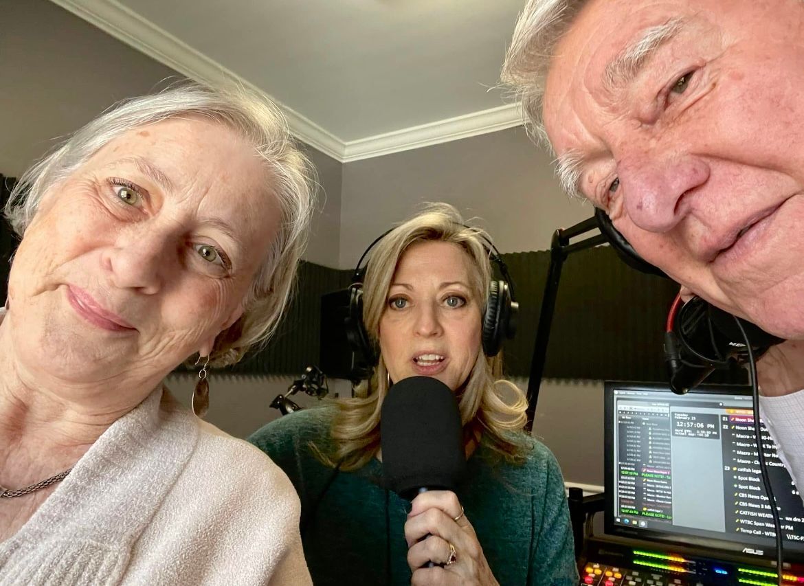 Three people in a podcast studio, one holding a microphone, with audio equipment and monitors visible.