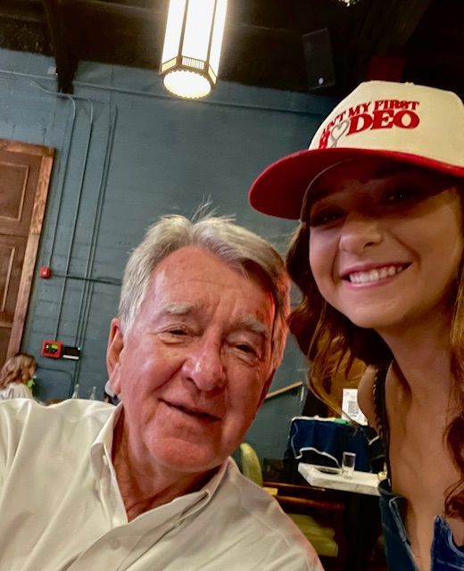 Two people smiling for a selfie indoors, one wearing a red-and-white cap.