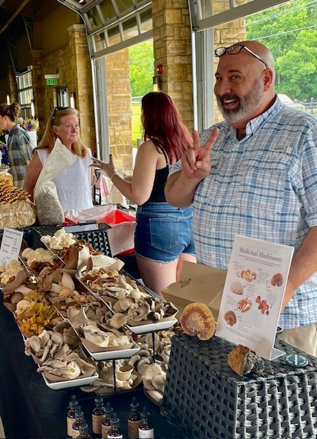 Vendor table with mushrooms and a smiling man making a peace sign at an indoor market.