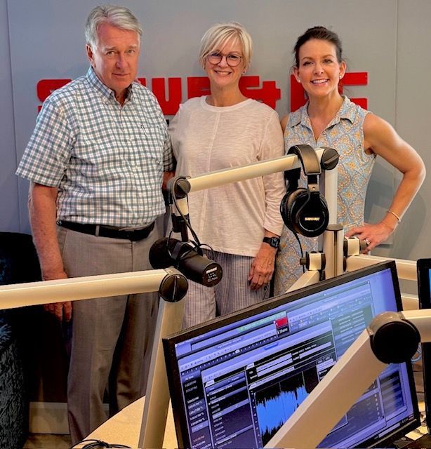 Three people standing in a radio studio behind microphones and computer monitors